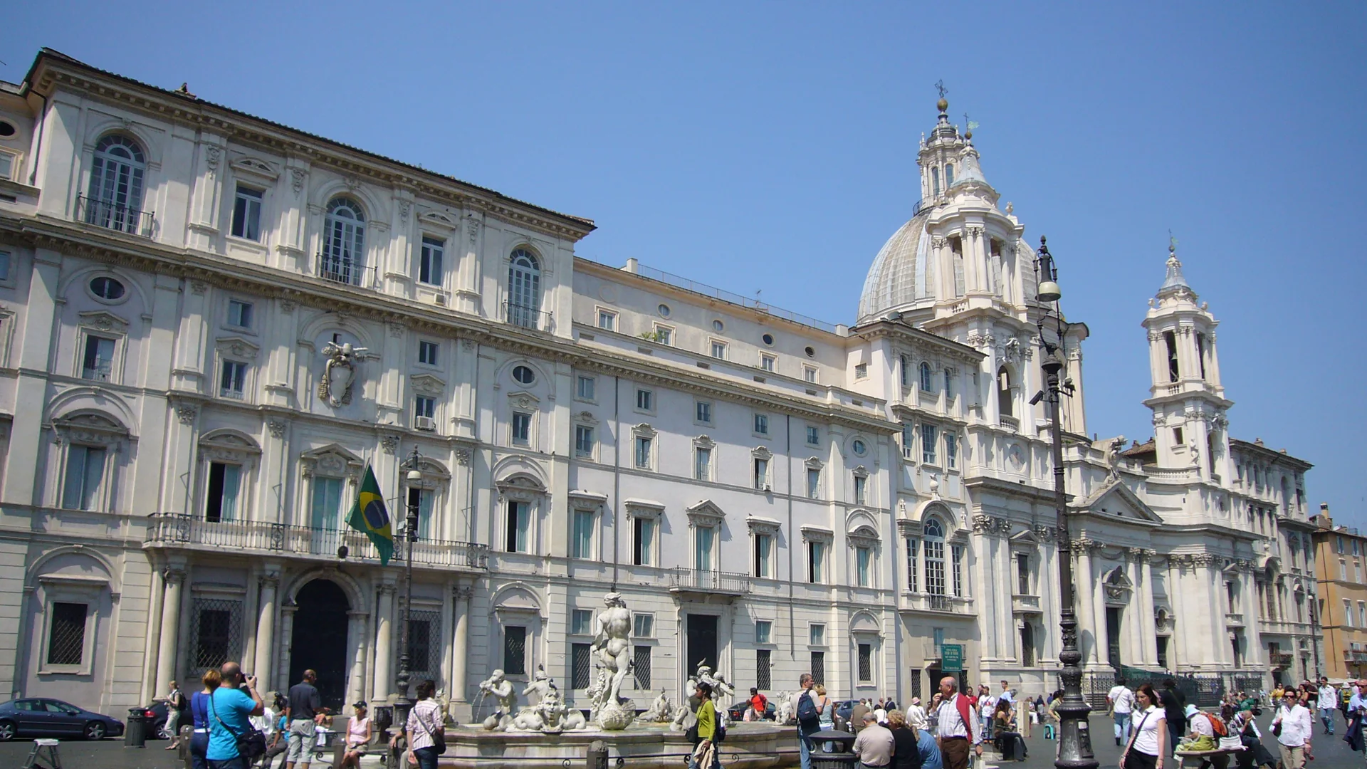 Krémová fasáda Palazzo Pamphilj na Piazza Navona, vedle ní se pod modrou oblohou tyčí kupole a zvonice Sant'Agnese in Agone.