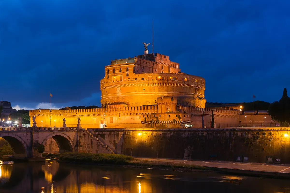 Castel Sant'Angelo badet i gyldent aftenlys ved Tiberen, en rund stenfæstning med en bronceengel på toppen og en buet bro i forgrunden